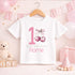Personalised 1st birthday girl outfit with pink number 1, cute cherry bow design, happy birthday text and custom name, shown on bib, vest, sleepsuit, t-shirt and white pyjamas.