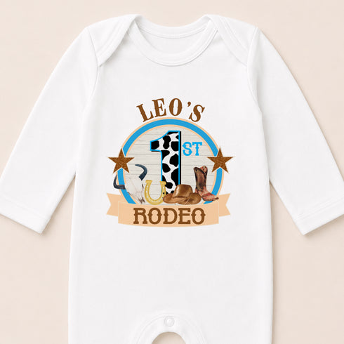A white onesie with a graphic design featuring a cow, a horse, and a cowboy hat, along with the text "LEO'S 1ST RODEO".