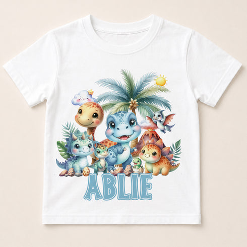 A white t-shirt with a colorful illustration of various cartoon-style dinosaurs and animals, including a smiling dinosaur and a palm tree. The word "ABLIE" is written below the illustration.