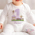 "An adorable baby wearing a personalized birthday onesie. The design showcases a large purple number '1,' flanked by whimsical fairies, a mushroom house, and a wooden fence surrounded by flowers. The text reads, 'HAPPY BIRTHDAY Florence,' celebrating a special little one's milestone!