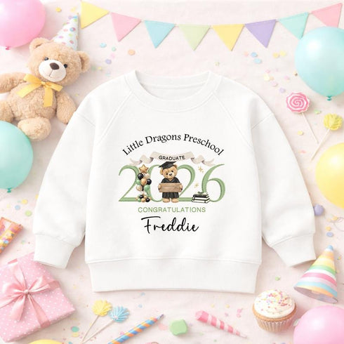 White children’s sweatshirt with a personalised green 2026 teddy bear nursery graduation design, displayed with colourful balloons, bunting and party decorations.