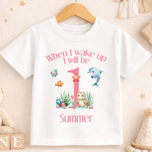 A white t-shirt with a printed design featuring a pink number one surrounded by various sea creatures, including a dolphin, starfish, and octopus. The text on the t-shirt reads "When I wake up I will be 1 Summer".