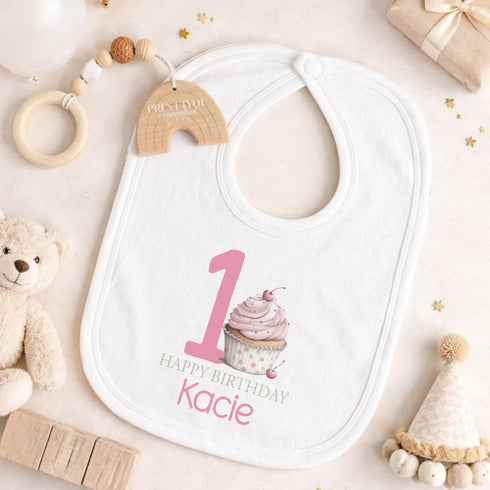 Personalised 1st birthday girl outfit with pink number 1, cute cupcake design, happy birthday text and custom name, shown on sleepsuit, t-shirt, vest, pyjamas and bib.