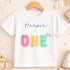 A white t-shirt with the text "Harper is ONE" printed on it, along with colorful lettering and floral designs.