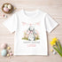 A white t-shirt with a bunny graphic and the text "Happy Easter Lottie" printed on it, along with Easter decorations such as a basket of eggs, flowers, and a banner.