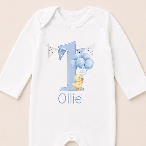 A white onesie with a blue number one and a yellow duck holding blue balloons, along with the name "Ollie" printed on it.