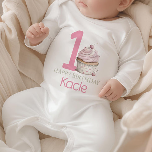Personalised 1st birthday girl outfit with pink number 1, cute cupcake design, happy birthday text and custom name, shown on sleepsuit, t-shirt, vest, pyjamas and bib.