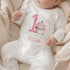 Personalised 1st birthday girl outfit with pink number 1, cute cupcake design, happy birthday text and custom name, shown on sleepsuit, t-shirt, vest, pyjamas and bib.