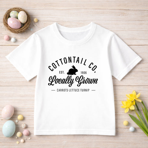A white t-shirt with text and a graphic of a rabbit, surrounded by Easter eggs, flowers, and other decorative items on a wooden surface.