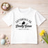 A white t-shirt with text and a graphic of a rabbit, surrounded by Easter eggs, flowers, and other decorative items on a wooden surface.