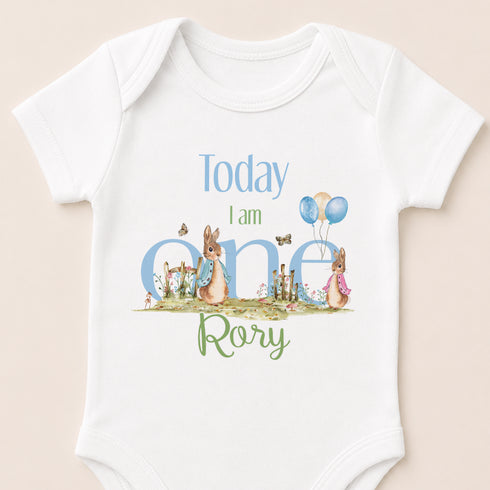A white baby onesie with a design featuring two rabbits and balloons, along with the text "Today I am one" and "Rory" printed on it.