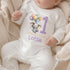 Personalised 1st birthday baby outfit with cute elephant, pastel balloons, purple number 1 and custom name, shown on bib, vest, sleepsuit, t-shirt and white pyjamas.