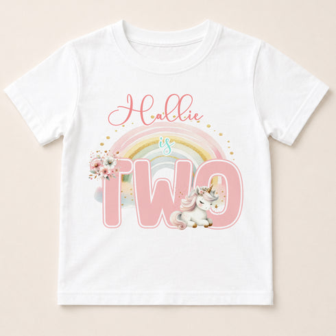 A white t-shirt with a pink unicorn and a rainbow graphic, along with the text "Hollie is two".