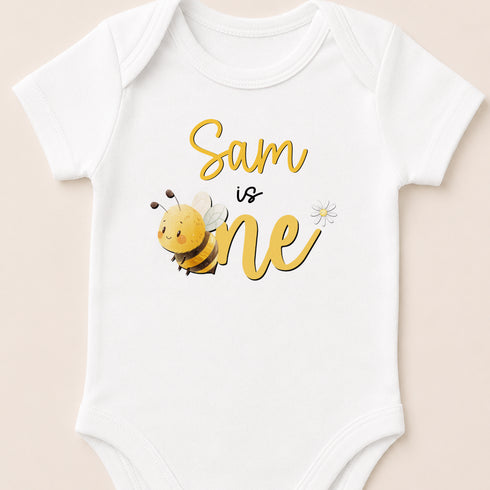 A white baby onesie with a yellow bee and the text "Sam is one" printed on it.
