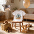 A white t-shirt with a cowboy-themed design, a balloon, a cowboy hat, and a pair of boots, all arranged on a wooden chair in a room.