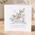 A greeting card featuring a mother rabbit and her baby bunny, surrounded by floral elements and a blue ribbon. The card has a personal message that reads "To my wonderful mummy, love from James" and is displayed on a wooden surface.