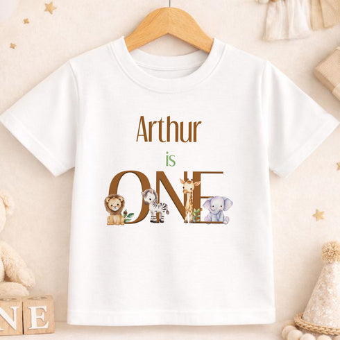 A white t-shirt with the text "Arthur is ONE" printed on it, featuring illustrations of various animals such as a lion, zebra, and elephant.