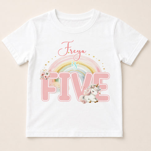 A white t-shirt with a pink unicorn and rainbow graphic, along with the text "Freya is FIVE" printed on it.