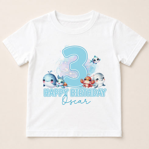 A white t-shirt with a graphic design featuring a large number 3, surrounded by cartoon-style illustrations of various sea creatures, and the text "Happy Birthday Oscar" below.