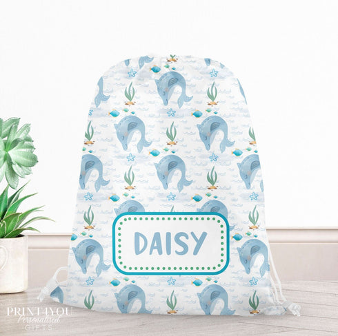 A white drawstring bag with a pattern of blue and green sea creatures, including a dolphin and a whale, printed on it. The bag has a name tag that says "DAISY". The bag is placed on a wooden surface next to a potted plant.