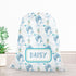 A white drawstring bag with a pattern of blue and green sea creatures, including a dolphin and a whale, printed on it. The bag has a name tag that says "DAISY". The bag is placed on a wooden surface next to a potted plant.