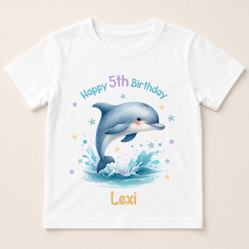 A white t-shirt with a printed design of a dolphin jumping out of the water, surrounded by stars and splashes of water. The text "Happy 5th Birthday Lexi" is displayed above the dolphin.