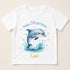 A white t-shirt with a printed design of a dolphin jumping out of the water, surrounded by stars and splashes of water. The text "Happy 5th Birthday Lexi" is displayed above the dolphin.