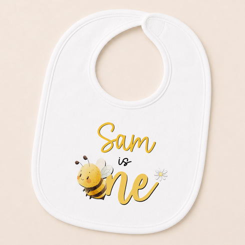 A white baby bib with a cartoon bee and the text "Sam is one" printed on it.