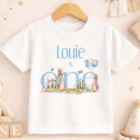 A white t-shirt with a personalized text and illustrations, including a rabbit and balloons, is displayed on a wooden hanger.