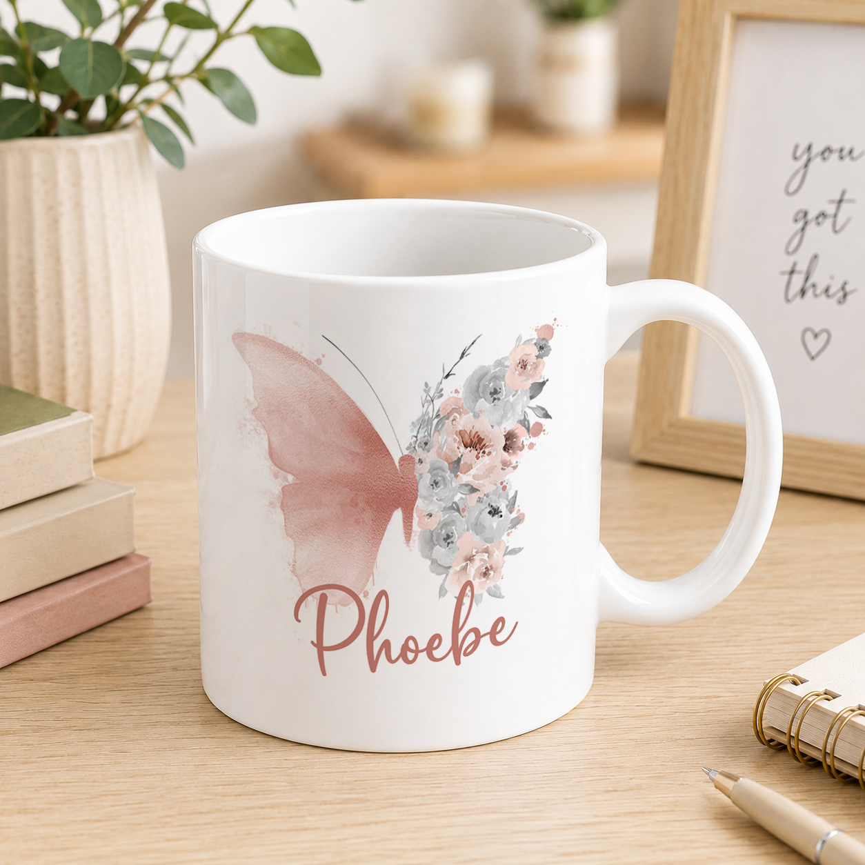 A white ceramic mug with a floral design and the name "Phoebe" printed on it, placed on a wooden surface next to some books and a plant.