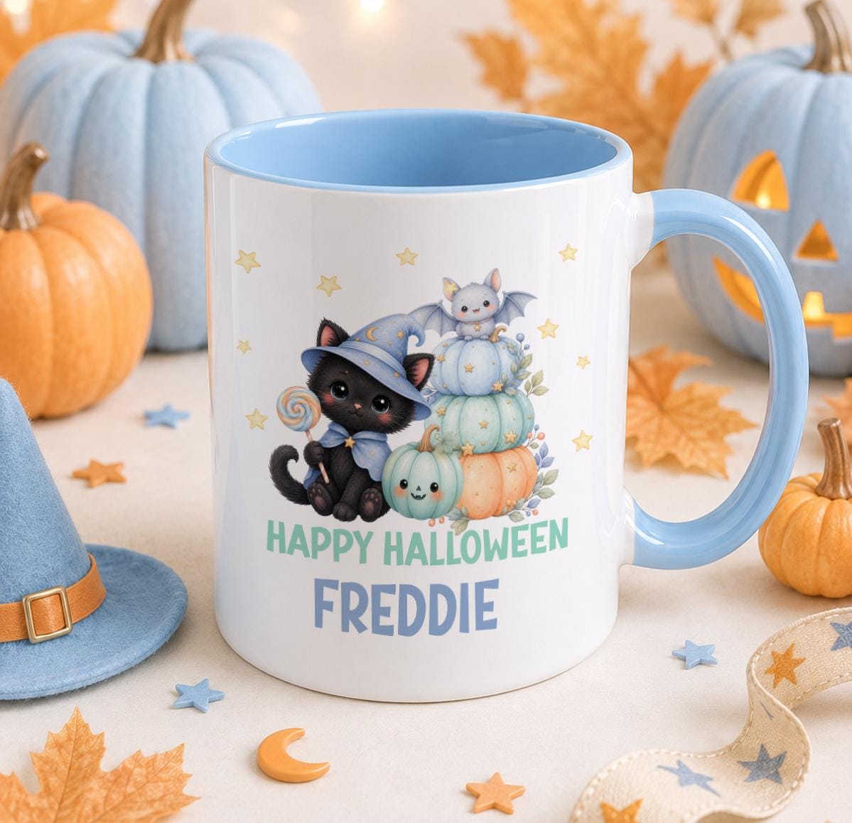 White ceramic mug with pale blue handle and inner, printed with a black kitten in a blue witch hat holding a lollipop, a baby bat and stacked pastel pumpkins, personalised with the name Freddie.