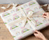 A person's hands are wrapping a gift with a white ribbon and a patterned wrapping paper featuring a unicorn, rainbows, and the name "Sophie".