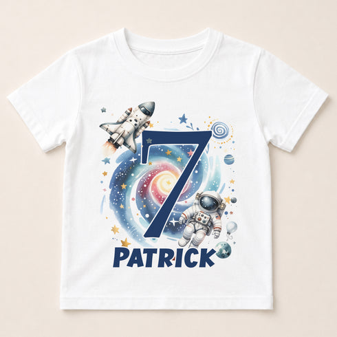 A white t-shirt with a space-themed design featuring a rocket ship, an astronaut, and a spiral galaxy. The number 7 is prominently displayed in the center, and the name "PATRICK" is written below the number.
