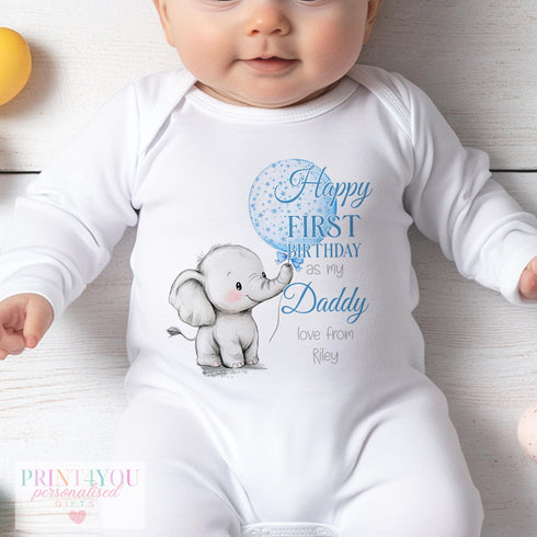 A baby dressed in a white sleepsuit featuring a charming design. The design includes a baby elephant holding a balloon. The text reads: "Happy FIRST BIRTHDAY as my Daddy love from Riley." The background displays a wooden surface and a yellow ball placed on the left. The onesie is from the brand "PRINT4YOU personalised GIFTS," showcasing their customisable products.