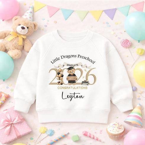 White children’s sweatshirt with a personalised gold 2026 teddy bear nursery graduation design, displayed on a colourful celebration background with balloons and bunting.
