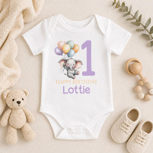 Personalised 1st birthday baby outfit with cute elephant, pastel balloons, purple number 1 and custom name, shown on bib, vest, sleepsuit, t-shirt and white pyjamas.