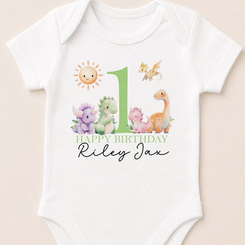 A white baby onesie with a green number 1 and various cartoon animal illustrations, including a sun, dragon, and unicorn. The text "Happy Birthday Riley" is displayed below the illustrations.