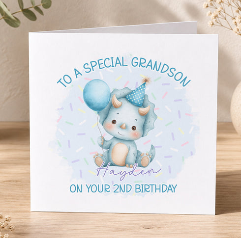 A birthday card with a cute illustration of a baby dinosaur wearing a party hat and holding a blue balloon, along with the text "To a special grandfather on your 2nd birthday".