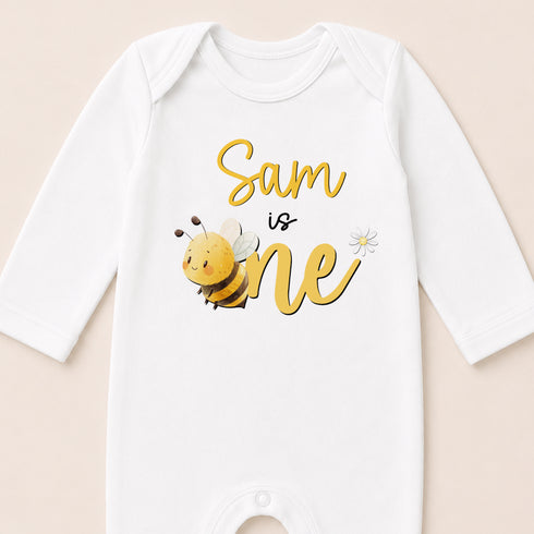 A white onesie with a yellow bee and the text "Sam is one" printed on it.