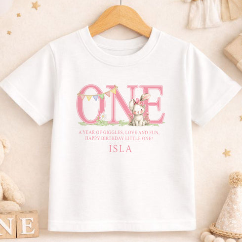 A white t-shirt with a pink text design that says "ONE" and features a bunny character. Below the text, there is a message that reads "A year of giggles, love and fun. Happy birthday little one! ISLA".