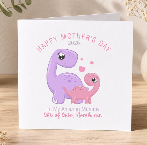 A greeting card with a cartoon illustration of a purple dinosaur and a pink dinosaur, along with the text "Happy Mother's Day 2026" and "To My Amazing Mummy lots of love, Norah xxx".