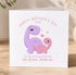 A greeting card with a cartoon illustration of a purple dinosaur and a pink dinosaur, along with the text "Happy Mother's Day 2026" and "To My Amazing Mummy lots of love, Norah xxx".