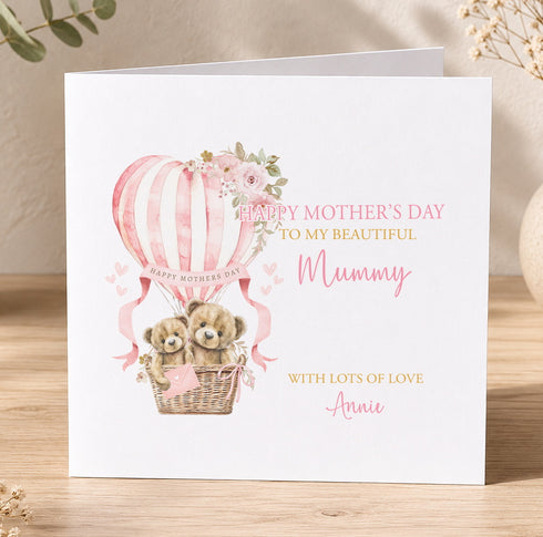 A white greeting card with a pink and white design featuring two teddy bears in a basket, an air balloon, and flowers. The card has a message that reads "Happy Mother's Day to my beautiful Mummy with lots of love Annie".