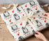A person's hands are wrapping a gift with a white ribbon and a patterned wrapping paper featuring penguins and red stars.