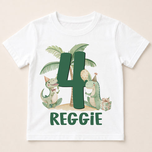 A white t-shirt with a graphic design featuring two dinosaurs, one larger than the other, and a palm tree. The larger dinosaur is wearing a party hat and holding a party balloon, while the smaller dinosaur is holding a gift box. The number "4" is prominently displayed in the center of the design. The text "REGGIE" is written below the image.