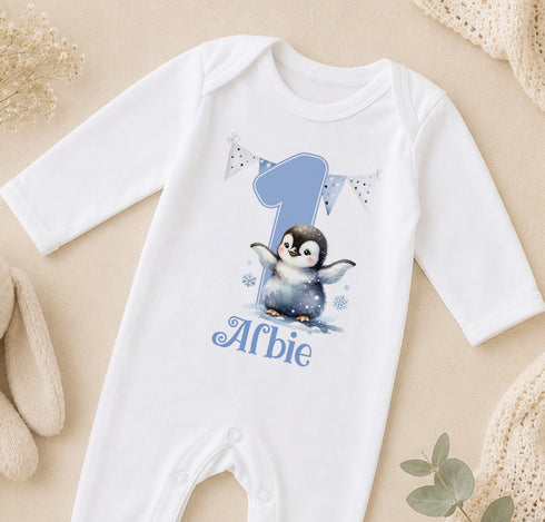 A white onesie with a penguin design and the number 1, along with the text "Alfie" printed on it, is displayed on a beige surface.
