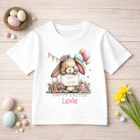 A white t-shirt with a cartoon rabbit holding a sign that says "Happy Easter" and "Lexie" printed on it, surrounded by Easter decorations such as balloons, flowers, and eggs.