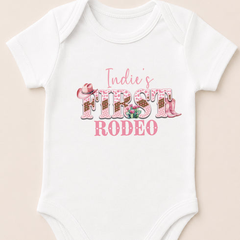 A white baby onesie with the text "Indie's FIRST RODEO" printed on it.
