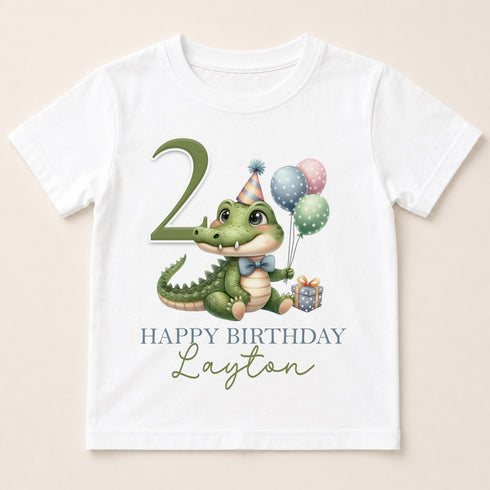 A white t-shirt with a cartoon image of a green alligator wearing a party hat and holding balloons, along with the text "Happy Birthday Layton" printed on it.