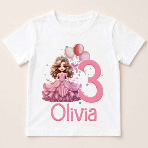 A white t-shirt with a cartoon image of a girl in a pink dress and a crown, along with balloons and the number 3. The text below the image says "Olivia".